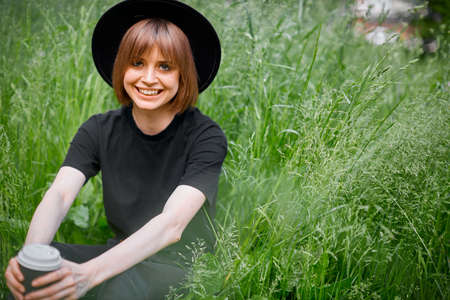 Young laughing girl in black and hat with a Cup of coffee in the green grass, eco-friendly active lifestyleの写真素材