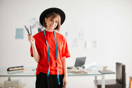 Young smiling girl fashion designer in hat standing with measuring tape and pencils in workshopの写真素材