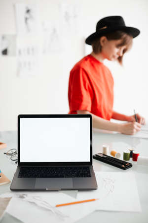 Young girl fashion designer in hat and laptop with empty screen in studio. Photo for ad educational courses or clothingの写真素材