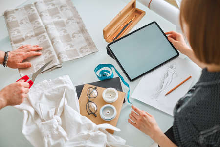 Pretty girl fashion designer works on tablet with empty screen in workshop. Photo for ad educational...