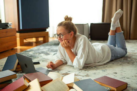 Cute teen girl student in casual clothes, glasses lying on floor at laptop with books. Back to...
