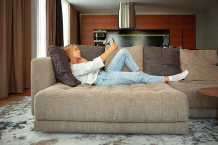 Pretty teen girl lying on sofa with phone talking via video chat or taking selfies. Cozy home environmentの写真素材