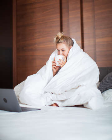 Cute teen girl under blanket in white bed in bedroom woken up and drink tea with laptop. Breakfast in bed, lazy morningの写真素材