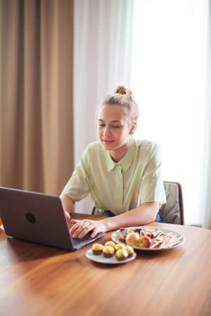 Teen girl sitting at the table with laptop watching TV series with delicious food. Cozy home environmentの写真素材