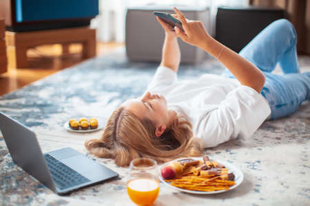 Teen girl lying on floor with laptop and phone scrolling the feed in social networks. Cozy home environmentの写真素材