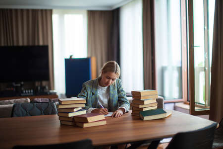 Young cute thoughtful teen girl student sitting with books at table in classroom or library. Back to...