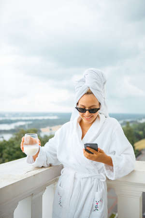 Smiling cute girl blogger in white robe, towel and sunglasses stands on balcony with phone and cup of milk. Lazy morningの写真素材