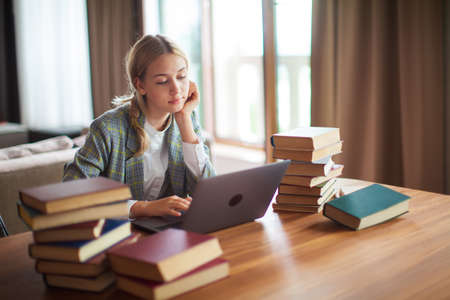 Young cute teen schoolgirl student sitting with books and working at laptop. Back to school. Exam...