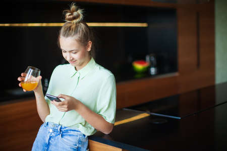 A young cute modern millennial girl with smartphone and a glass of juice at cozy homeの写真素材