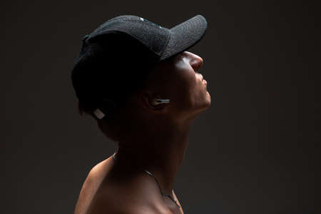 Cool young guy in cap and in a wireless earphone listen to song without shirt on black background....