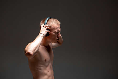 Cool young guy in wireless headphones with a naked torso listen to music in studio isolated on black backgroundの写真素材