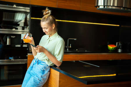 A young cute modern girl with smartphone and a glass of juice is relaxing in a cozy kitchen