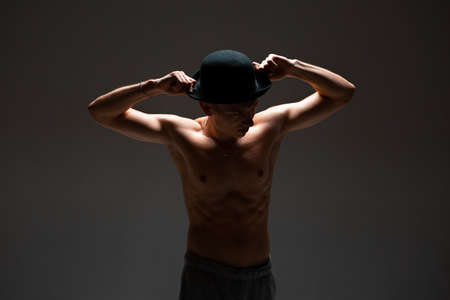 Stylish young guy actor in hat stands without shirt in studio isolated on black backgroundの写真素材