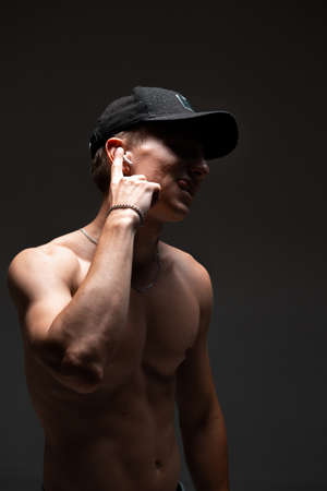 Cool young guy in cap and in wireless earphone listen to song with naked torso on black background. Music posterの写真素材