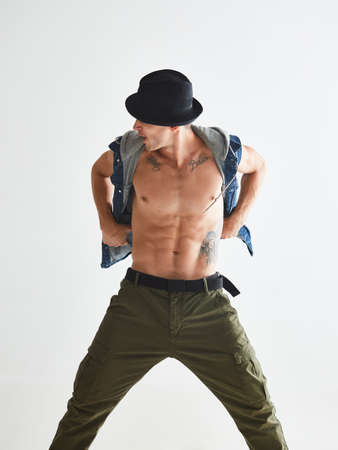 Cool young guy dancer in hat with naked torso standing in studio isolated on white background. Break...