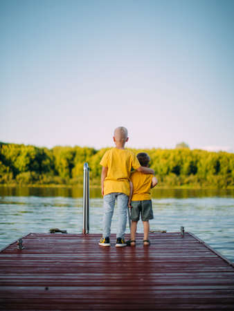 Cool kids brothers hug each other and look at the river staying on dock. Back viewの写真素材