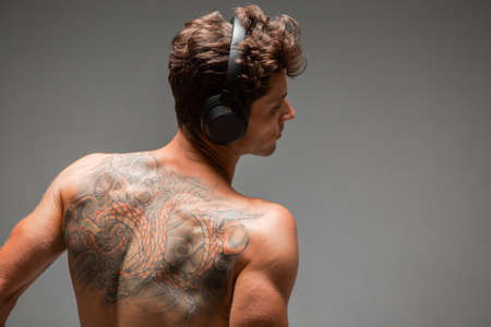 Cool young guy in wireless headphone listen to song with naked back isolated on gray background. Body with tattooの写真素材