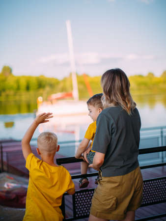 Young mother, cool kids boys stand on dock, look at the sailboat. Back view. Summer photography...