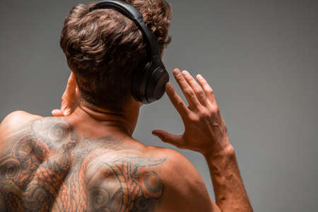 Cool young guy in wireless headphone listen to song with naked back isolated on gray background. Body with tattooの写真素材