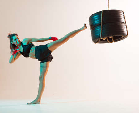 Female fighter practicing kicks using tires in studio isolated on beige background in neon. Mixed martial arts posterの写真素材