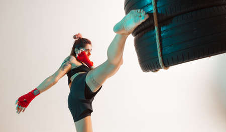 Cool girl fighter practicing kicks using tires in studio isolated on white background in neon. Mixed martial arts posterの写真素材
