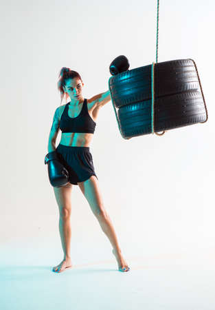 Boxer female fighter in boxing gloves posing in confident stance near punching bag of tires. Mixed martial arts posterの写真素材