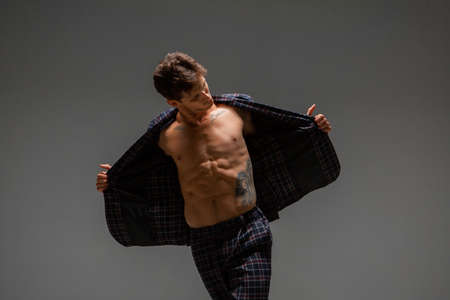 Handsome young guy in a wide-open jacket with naked torso stands in studio isolated on gray background. Body with tattooの写真素材