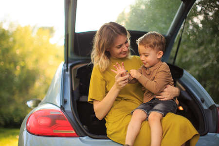 Beautiful young mother and little son travelers sit in trunk of car on road. Photo about family travelの写真素材