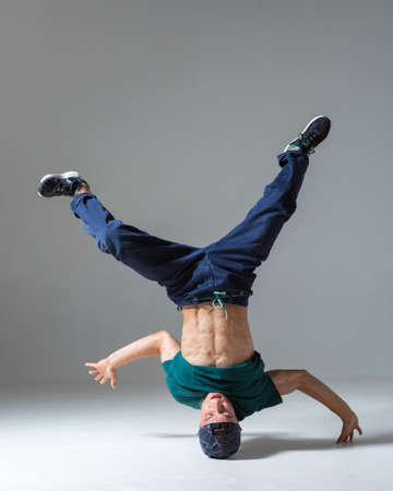 Stylish guy dancer dancing on head breakdance in studio isolated on gray background. Breakdance...