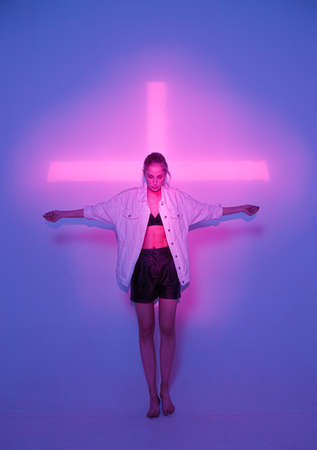 Cool young girl standing in violet neon light with pink cross in studio. Trendy poster for partyの写真素材