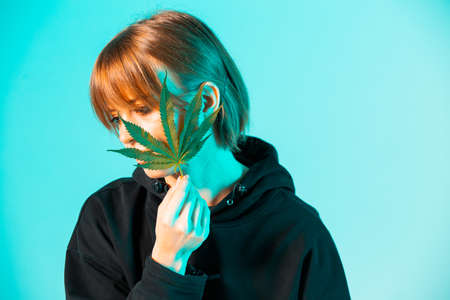 Portrait of young girl covering her face by hemp leaf in blue neon light