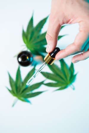 Pipette with CBD oil in male hand on hemp leaves and jar background, close up top view photo for ad...