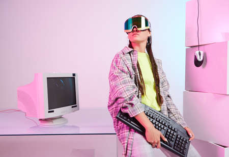 Stylish young female in goggles with old computer monitor, mouse, keyboard on workplace in office....