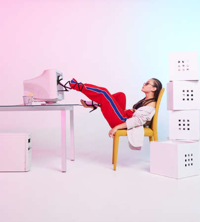 Stylish female office worker with obsolete computer and boxes sitting with feet on desk on...