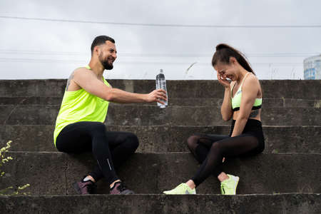 Athletic joyful young couple in stylish sportswear with bottle of water having break after workout in urban on stairsの写真素材
