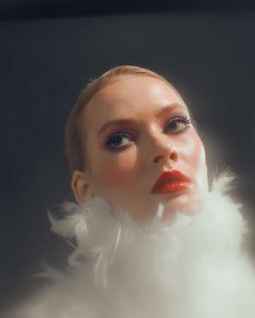 Romantic stylish young female model with white feathers collar, long lashes red lips, close up studio portraitの写真素材