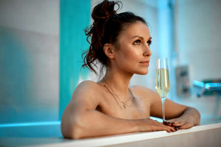 Dreamy young 30s female with champagne glass taking bath resting in bathroom in hotel or at homeの写真素材