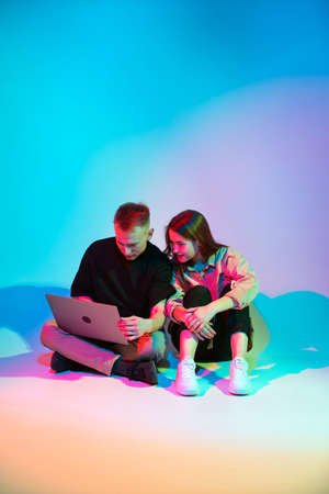 Modern young couple with laptop sitting on floor in colorful neon studio light working, playing or...