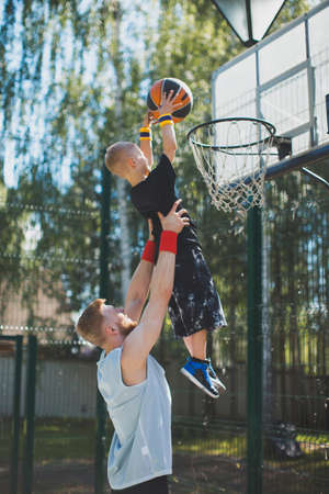 Young sportive father with son in hands playing basketball. Kid boy throwing ball in basketball hoop outdoorsの写真素材