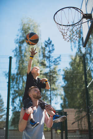 Dad and son playing basketball outdoors. Basketball player kid boy on fathers shoulders throw ball...