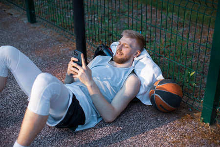 Cool basketball player sportive young guy lying relaxing with phone after workout outdoors on sports courtの写真素材