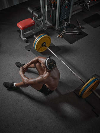 Athletic african american man with naked torso rest sitting on floor with barbell on training in...