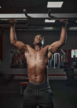 Athletic african american man with naked torso doing chin-ups or pull ups exercising on training in gymの写真素材