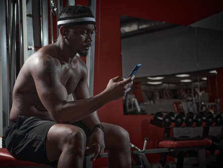 African american athletic man with phone using sports app for exercising and gym workout sitting on sport bench. High quality photoの写真素材