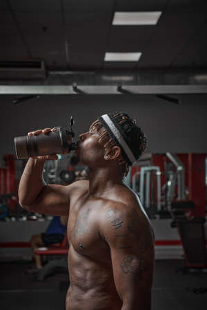 Sportive african american man with naked torso drink water or sports nutrition after training in gymの写真素材