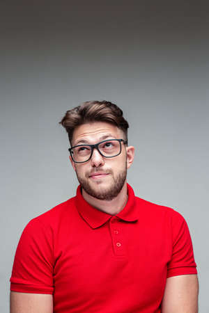 Pensive modern young man in glasses looking up on empty space wrinkling, studio portrait on gray...