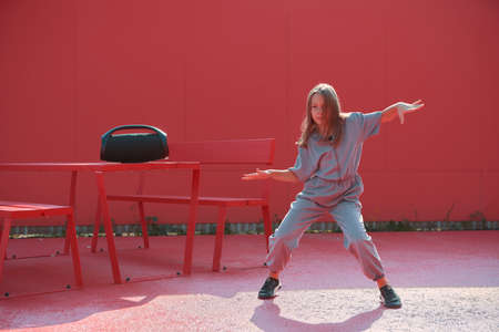 Cool child girl dancing with wireless outdoor speaker outside on red background. Dance school...
