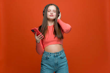 Modern young girl in wireless headphones enjoying sound listening music using mobile phone apps on orange backgroundの写真素材