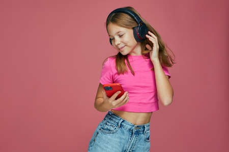Carefree cute little girl child in headphones dancing listening to music using phone apps on pink studio backgroundの写真素材