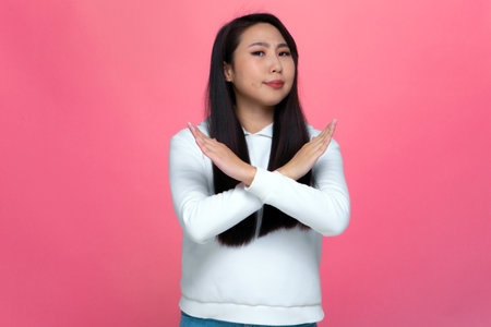 Stop gesture. Serious asian girl crossing hands, gesturing, prohibiting, restrict, reject, say no on pink backgroundの写真素材